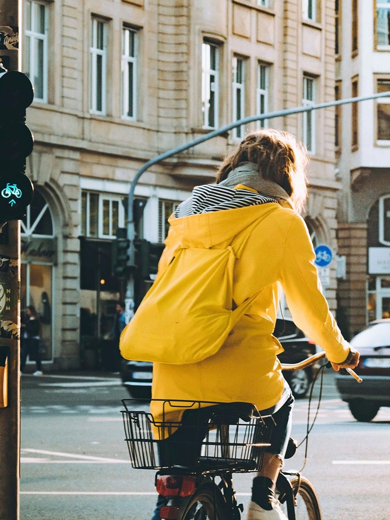 Frau mit gelbem Rucksack und gelber Jacke auf einem Fahrrad an einer Fahrradampel inmitten von traditionell aussehenden Häsuerblocks