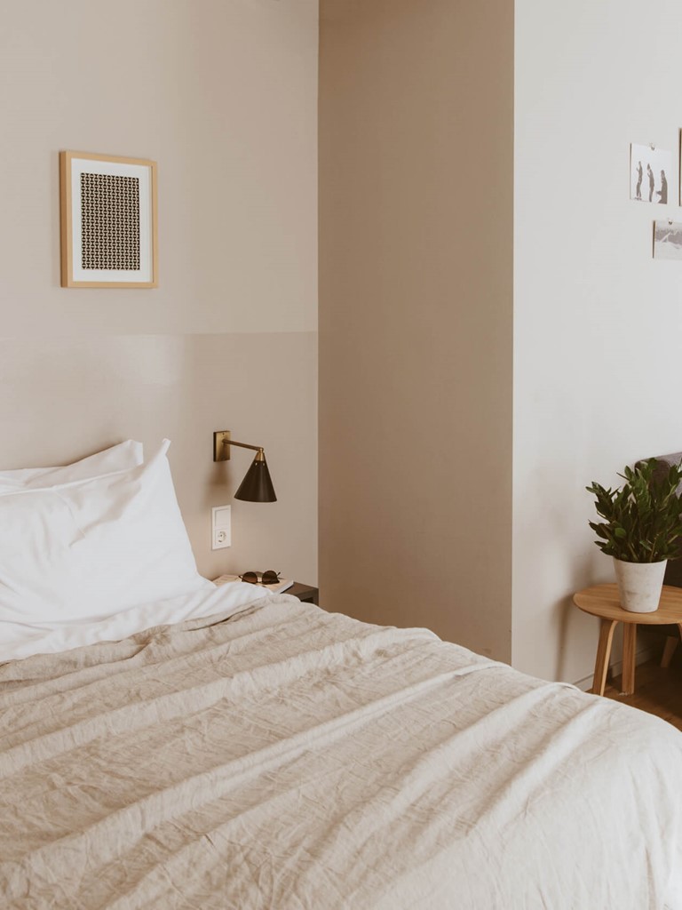 Rechts im Anschnitt ein hell bezogenes Doppelbett, ein graues Sofa mit farbig abgestimmten Kissen an der Wand, rechts dahinter im Anschnitt eine graue Küchenzeile.