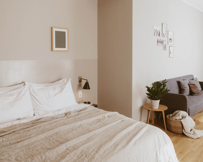 Rechts im Anschnitt ein hell bezogenes Doppelbett, ein graues Sofa mit farbig abgestimmten Kissen an der Wand, rechts dahinter im Anschnitt eine graue Küchenzeile.