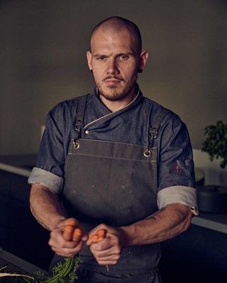 Man with bald head and beard in a kitchen dressed in a denim shirt and grey apron. He is breaking carrots apart with his hands.
