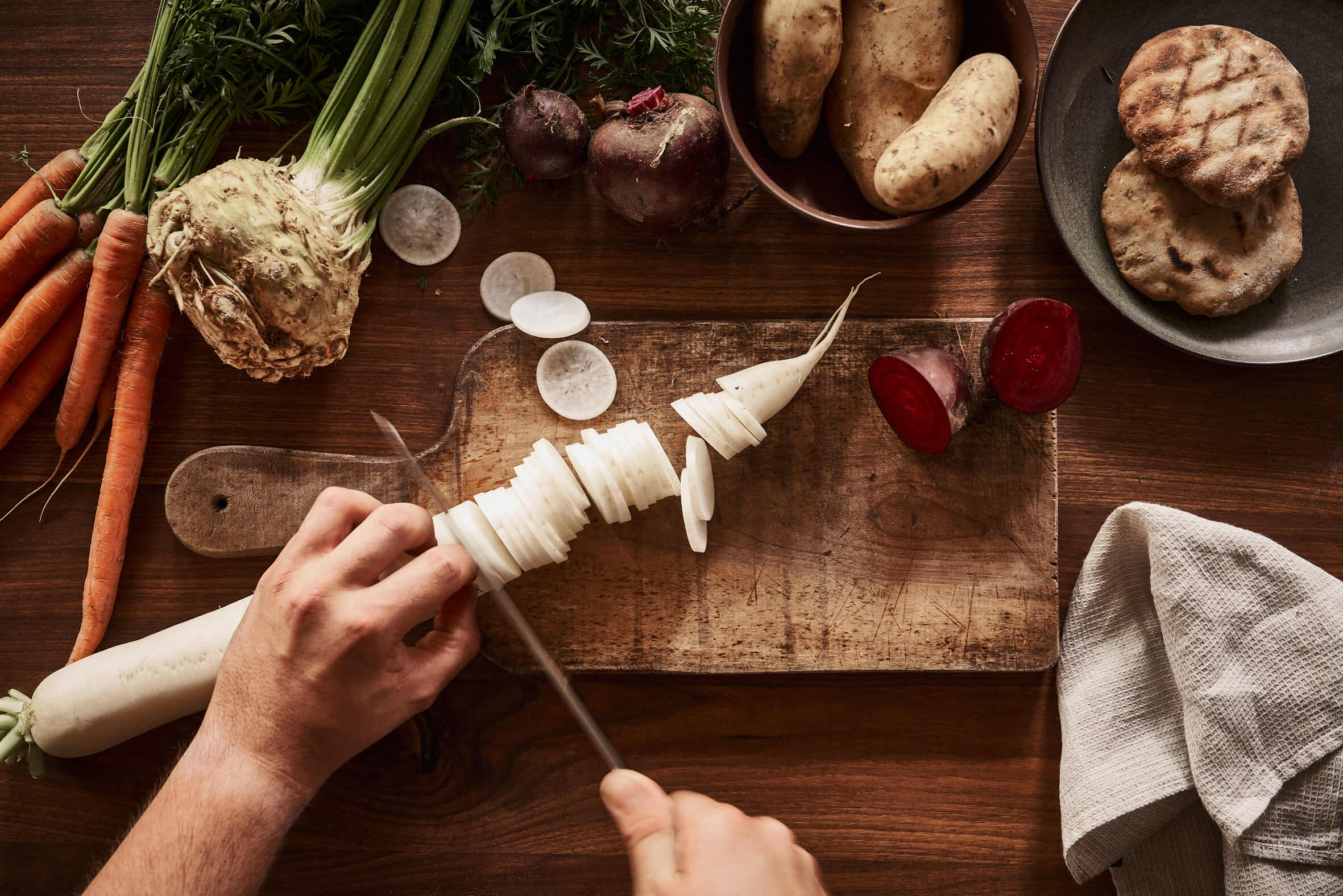 Hands of a person slicing a white radish on a wooden chopping board. Around the board are carrots, celeriac, beetroot and potatoes. 