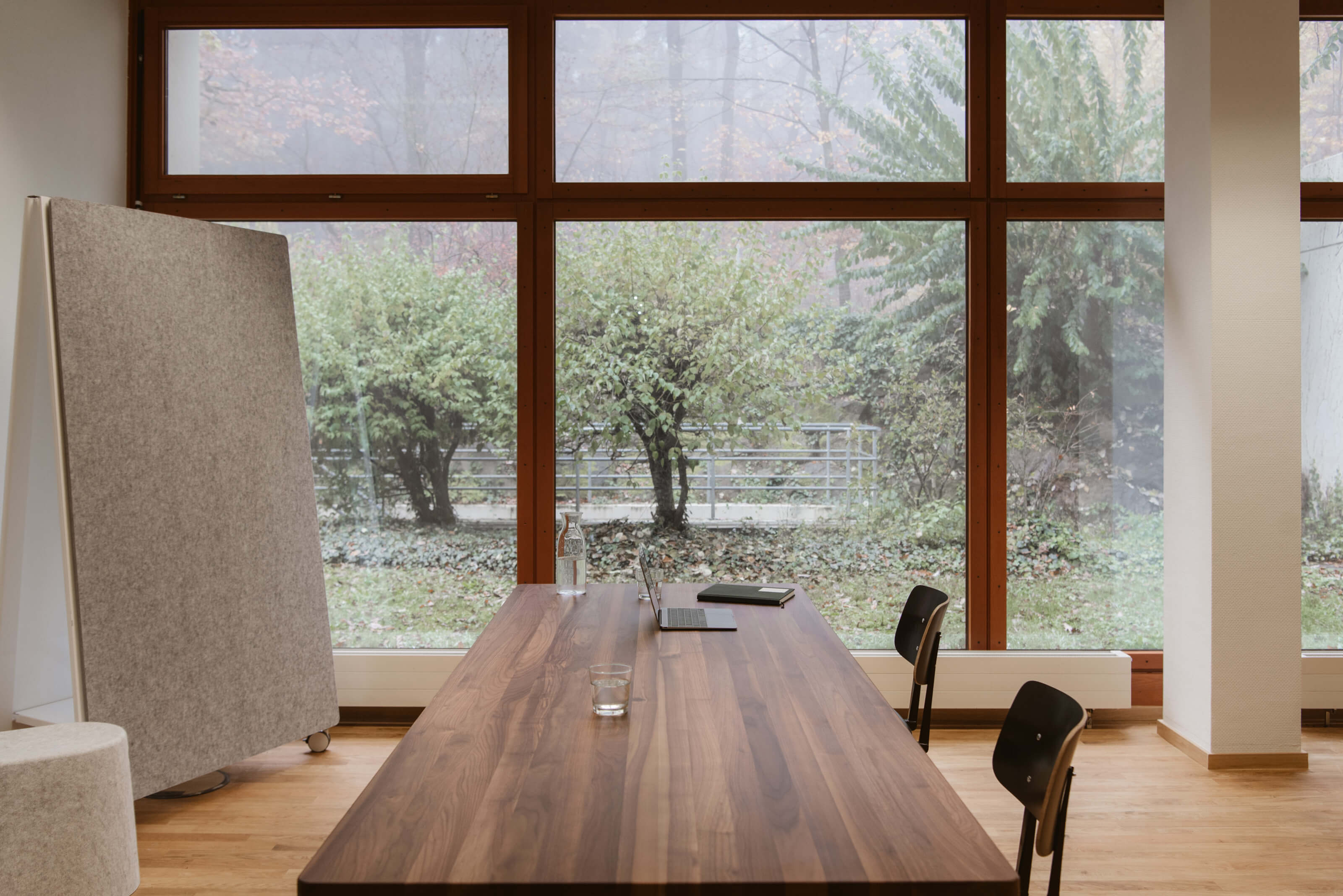 Konferenzraum in der Totale. Mittig ein Holztisch, seitlich daran zwei schwarze Stühlen. Links steht eine Pinnwand, frontal bodentiefe Fenster mit Blick ins Grüne.
