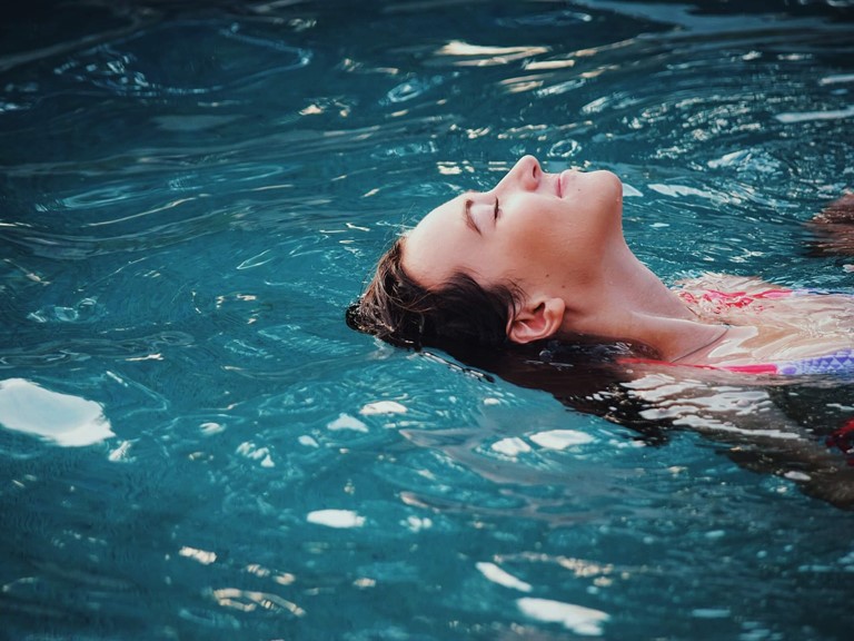 A dark-haired woman is floating in a pool on her back with her eyes closed, she is smiling.