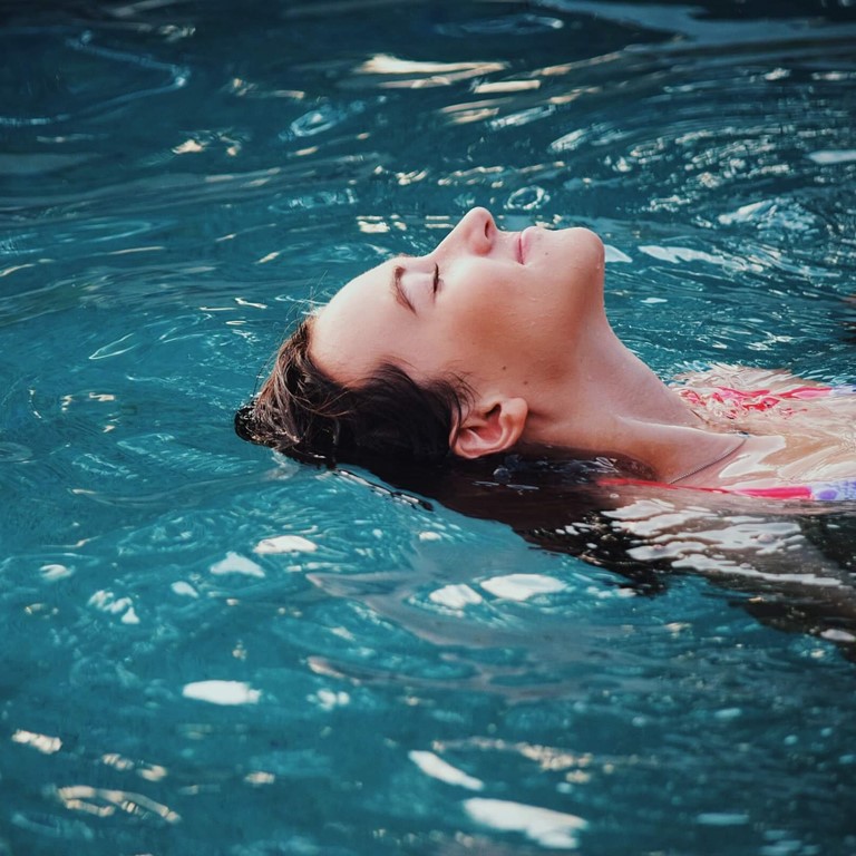 A dark-haired woman is floating in a pool on her back with her eyes closed, she is smiling.