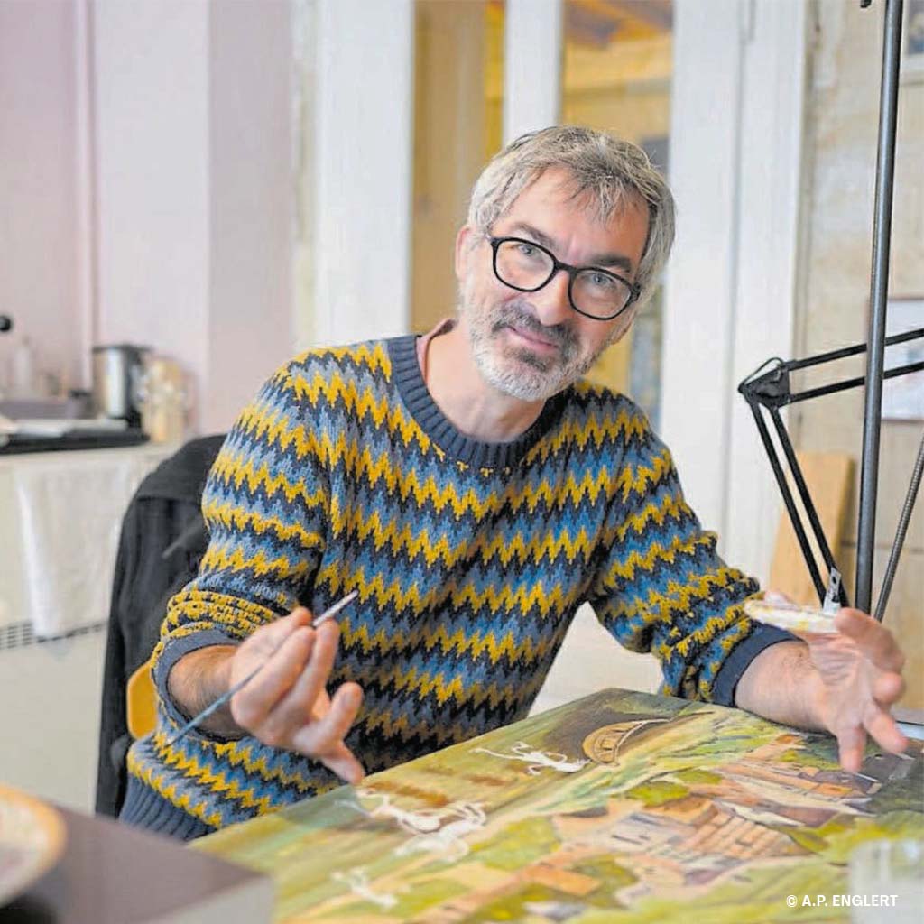 Sylvain Mérot, a smiling man with short grey hair, beard and glasses, draws on a canvas.