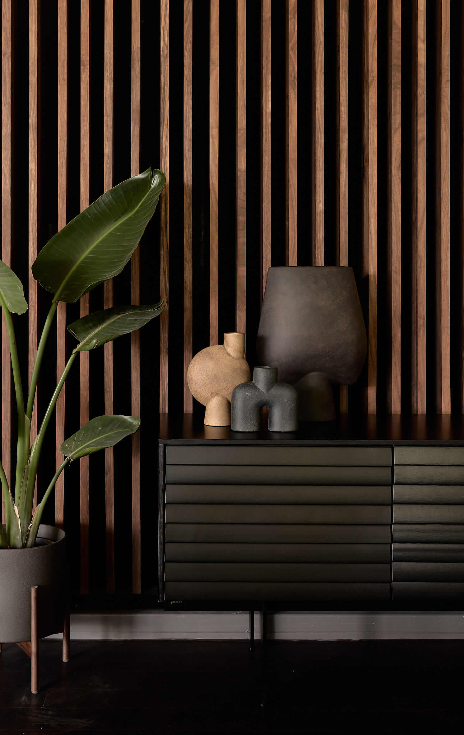 Moderne Vasen auf einem dunklen Sideboard vor einem Sichtschutz aus Holzelementen, links daneben eine Topfpflanze.