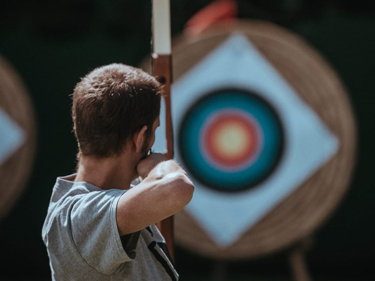Man in grey T-shirt with bow and arrow, arm at the ready, aiming at a target 