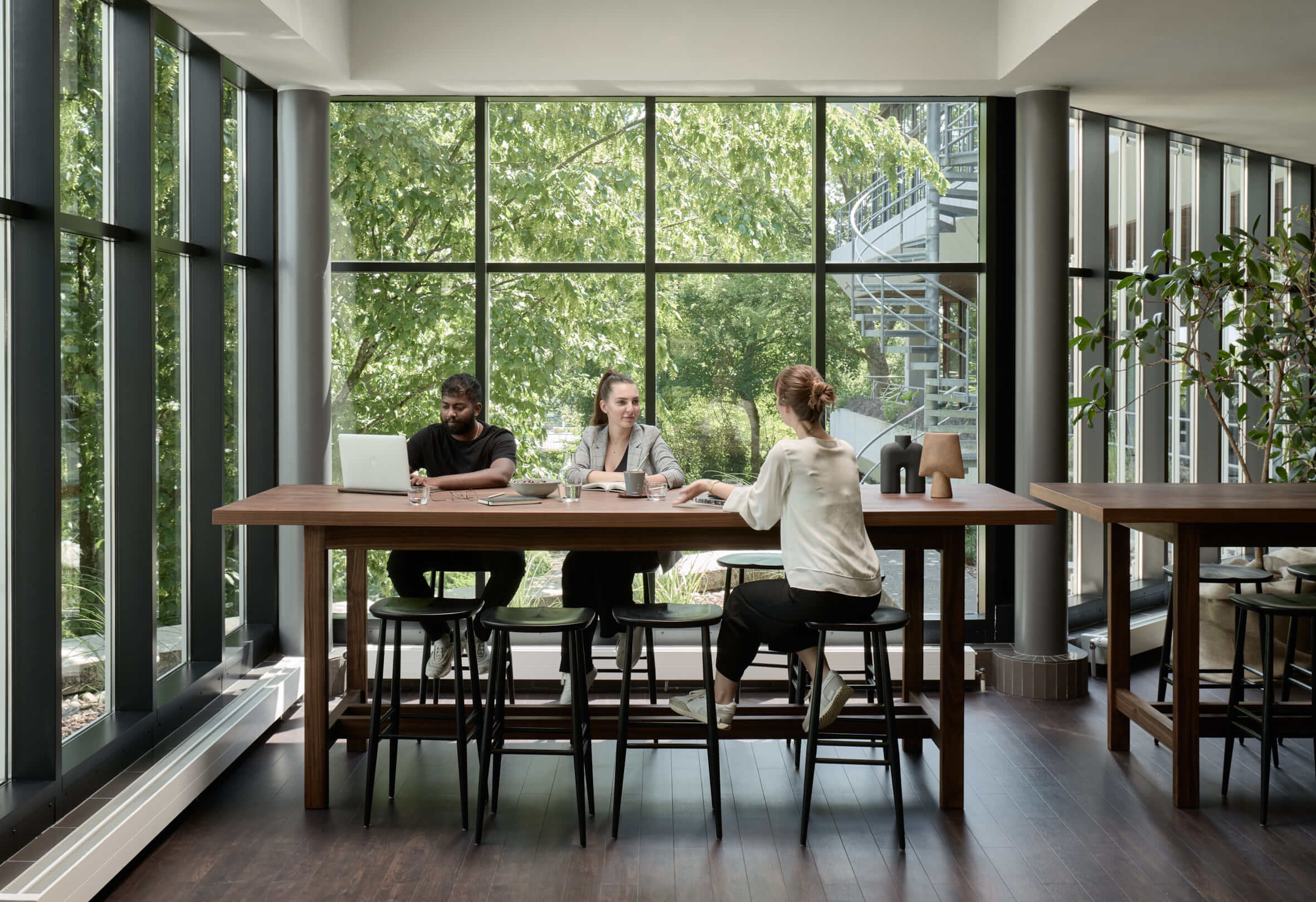 Ein Mann und zwei junge Frauen sitzen auf schwarzen Barhockern an einem hohen Holztisch und beraten sich. Durch die gläsernen Wände sind Pflanzen zu sehen. 