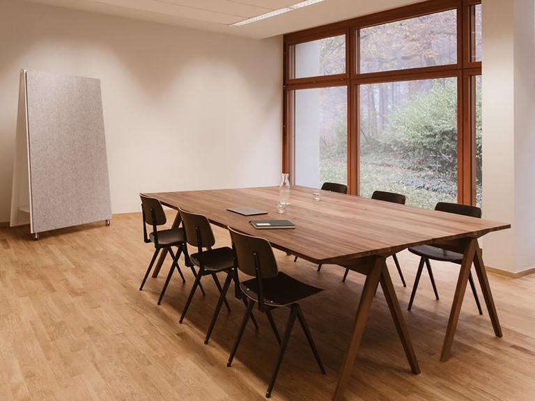 Meeting room with light wooden floor. In the middle, a dark wooden table with six black chairs around it. On the left is a large, mobile pin board, on the right you have a view of the green through floor-to-ceiling windows.