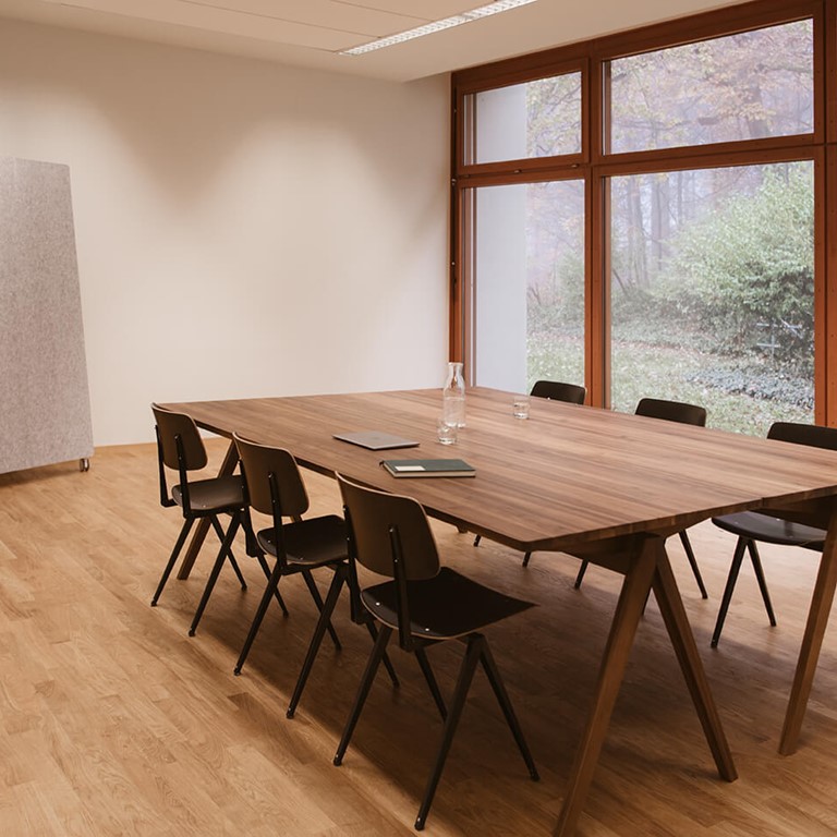 Meeting room with light wooden floor. In the middle, a dark wooden table with six black chairs around it. On the left is a large, mobile pin board, on the right you have a view of the green through floor-to-ceiling windows.
