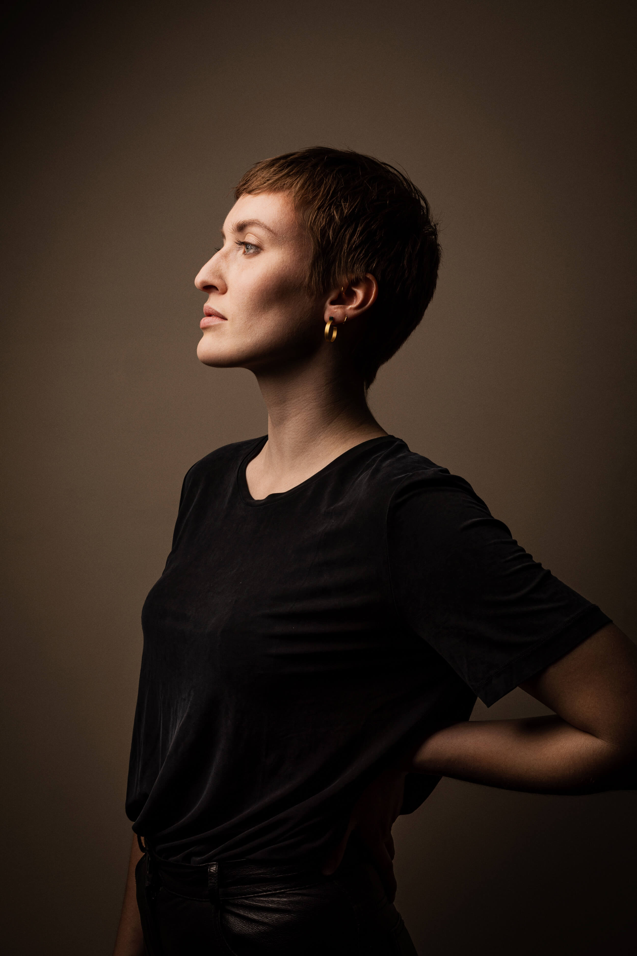 Eine Frau mit markantem Gesicht und kurzem, braunen Haar in einem schwarzem T-Shirt steht seitlich vor einem hellbraunen Hintergrund. Ihre rechte Hand hat sie leicht in den Rücken gestemmt.