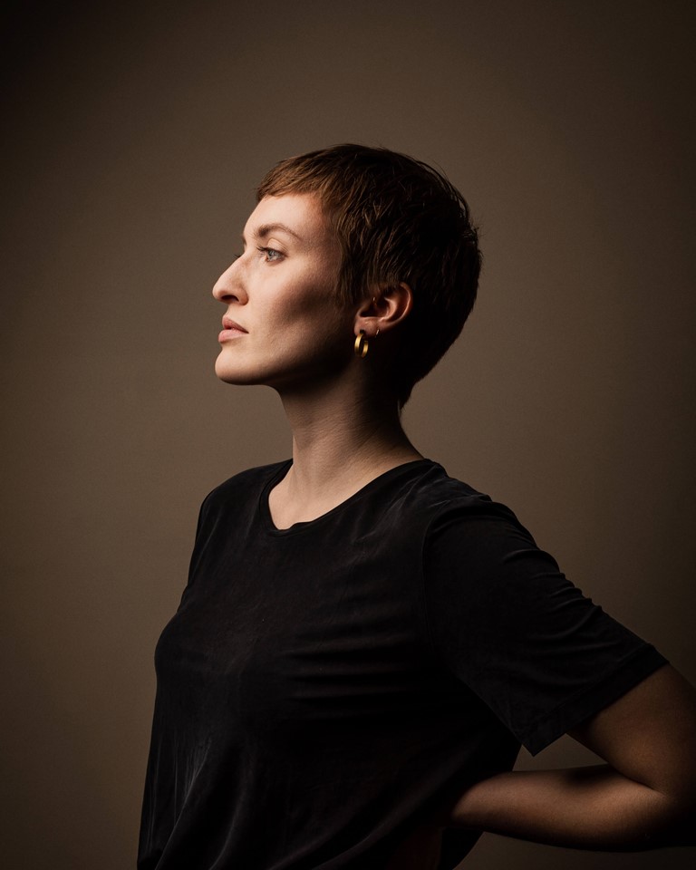 A woman with a striking face and short brown hair in a black T-shirt stands sideways in front of a light brown background. She has her right hand lightly resting on her back.