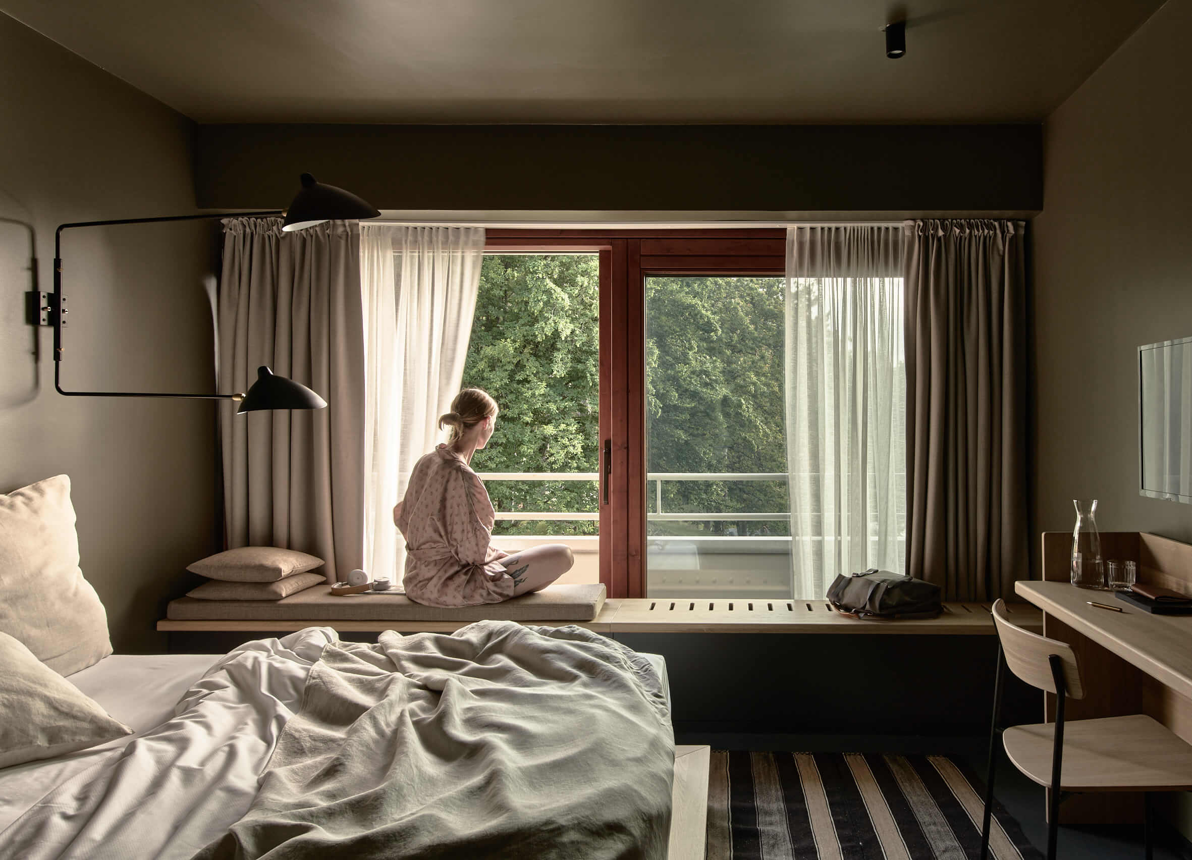 Woman in rose-coloured pyjamas sits on a bench by a large, open window and looks out. The view is from a modern but simply furnished room. 