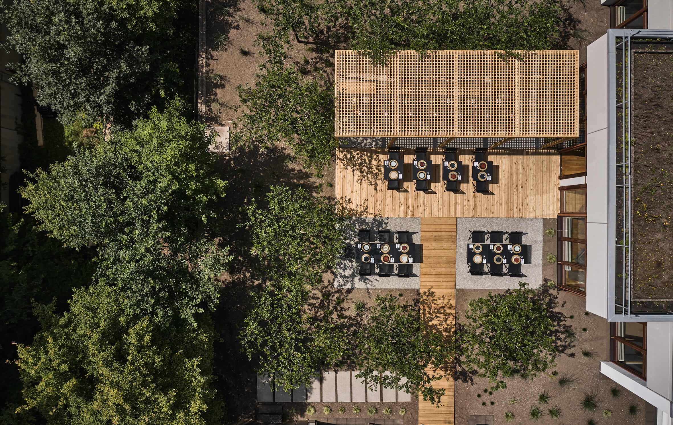 Luftaufnahme einer Terrasse mit gedeckten Tischen und Stühlen, eine hölzerne Pergola und viele Bäume daneben, im Anschnitt rechts ein Gebäude.