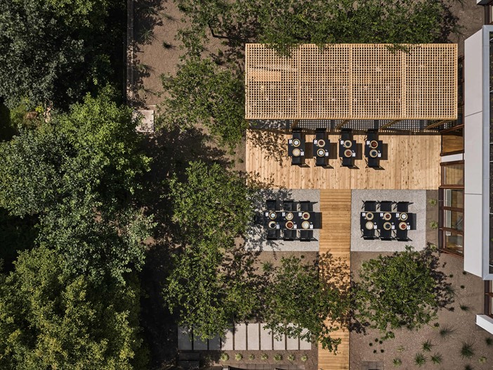 Aerial view of a terrace with laid tables and chairs, a wooden pergola and many trees next to it, in the section on the right a building.