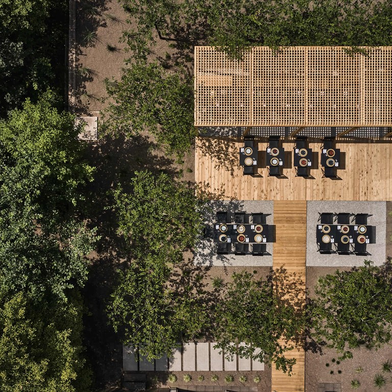 Aerial view of a terrace with laid tables and chairs, a wooden pergola and many trees next to it, in the section on the right a building.