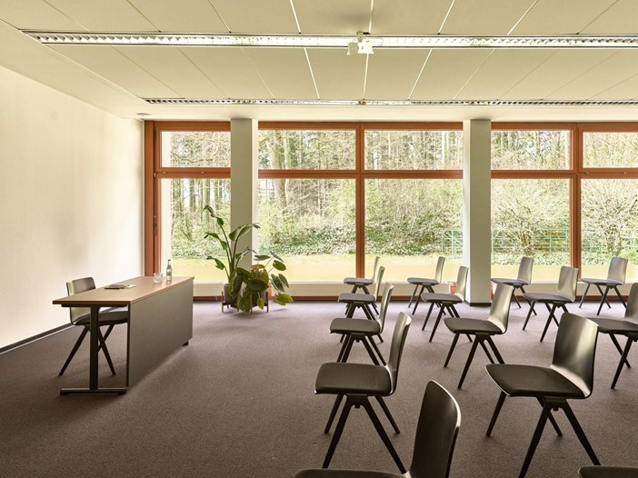 A conference room with floor-to-ceiling windows and a view of the greenery. There are a few chairs in the conference room. These are orientated towards a table.