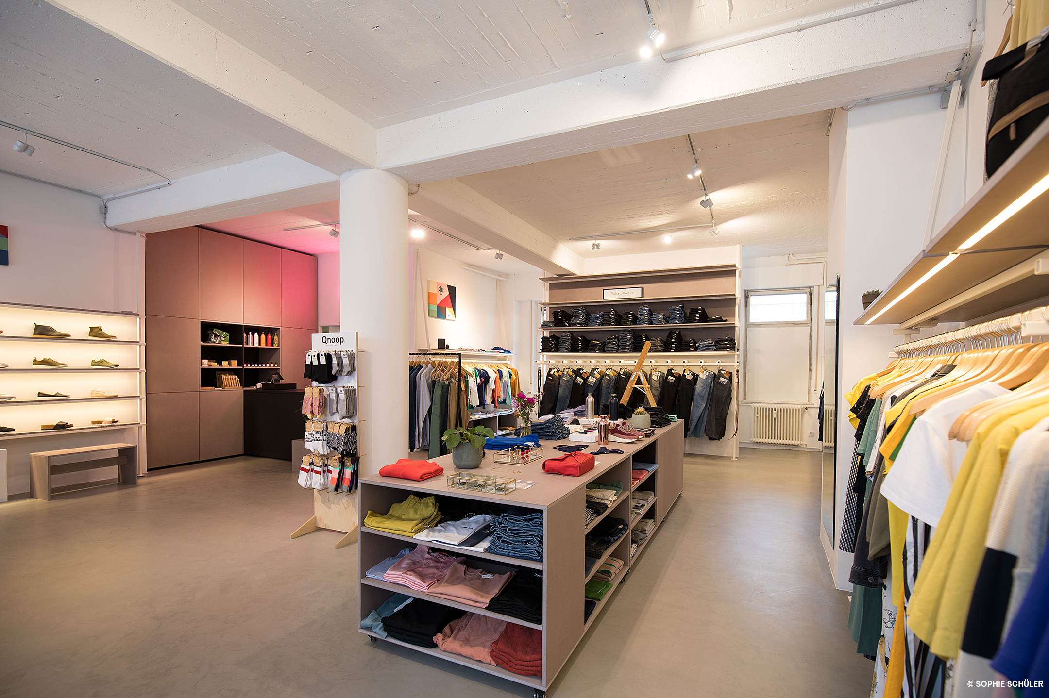 A minimalistically furnished clothing shop with shelves in light tones and colourful clothes displayed in and on the racks and hangers. 