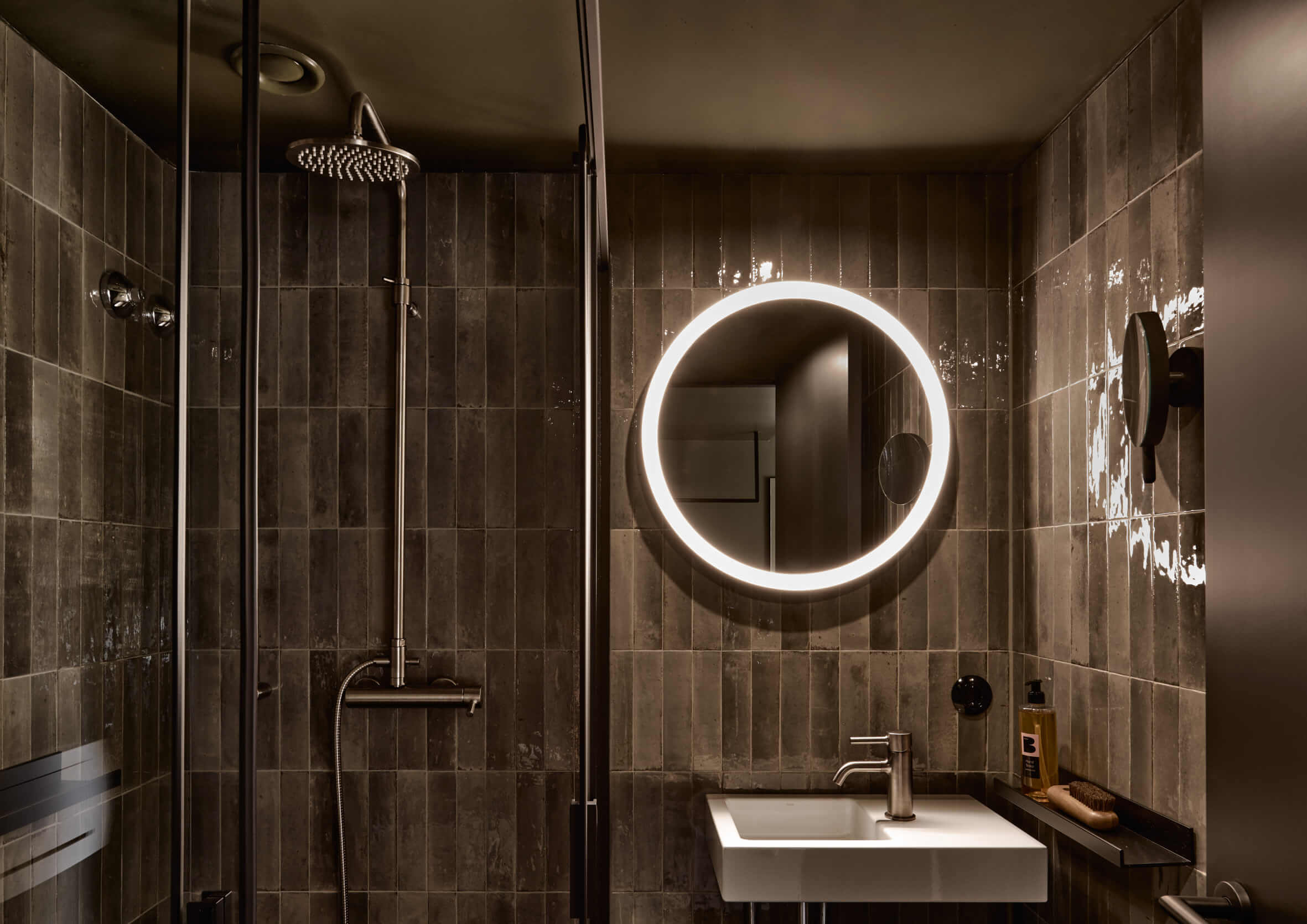 Modern tiled bathroom with rain shower, round illuminated mirror in the centre, white angular washbasin and accessories below.