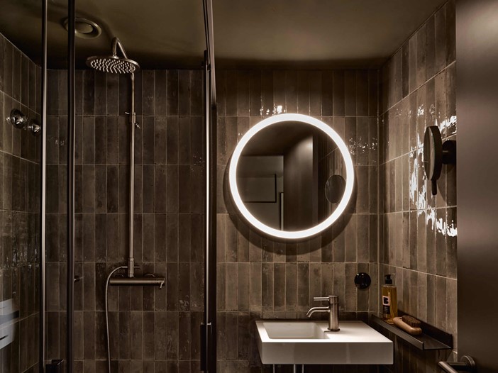 Modern gefliestes Badezimmer mit Regendusche, rundem, beleuchteten Spiegel in der Mitte, darunter ein weißes, eckiges Waschbecken und Accessoires.