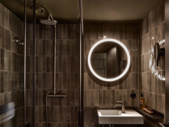 Modern gefliestes Badezimmer mit Regendusche, rundem, beleuchteten Spiegel in der Mitte, darunter ein weißes, eckiges Waschbecken und Accessoires.