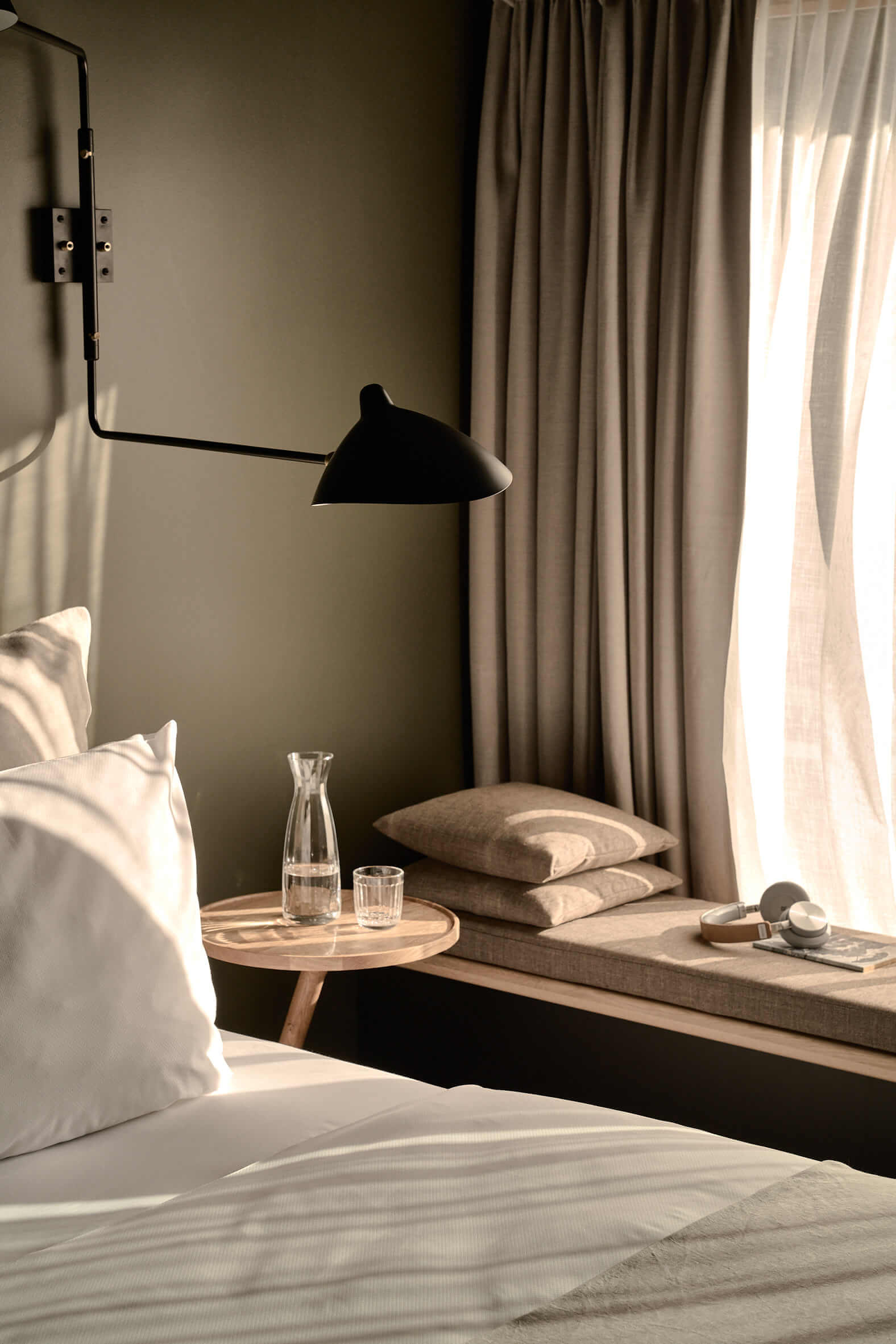 Cut of a brightly made-up bed, next to it a small wooden table with a water jug and glass, a modern, black wall lamp above it. To the right of the window, an upholstered seating corner with accessories.