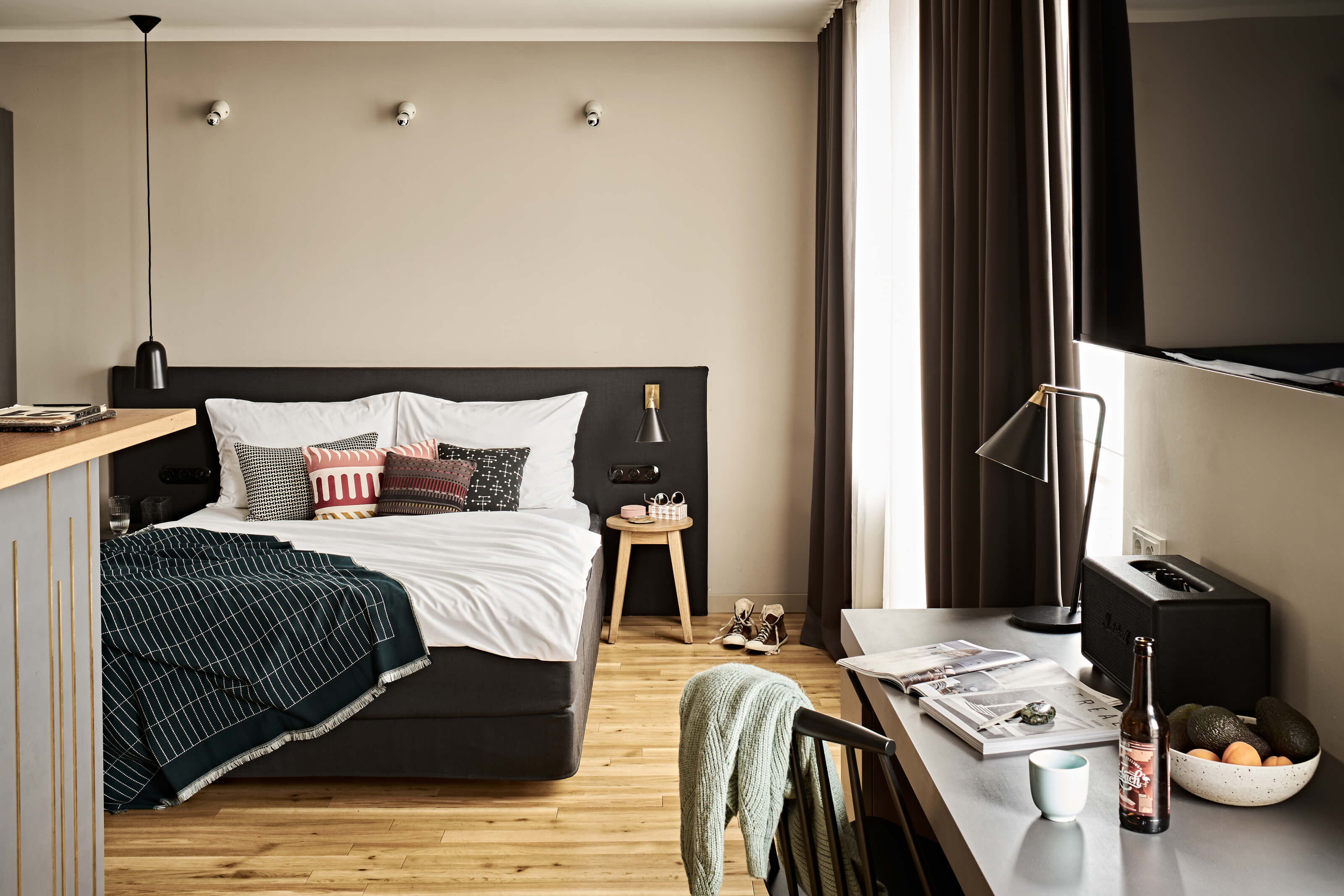 A comfortable-looking double bed with colourful pillows, a counter on the left, a large window with white and grey net curtains on the right, a desk with a chair against the wall, a flat-screen TV above it. 