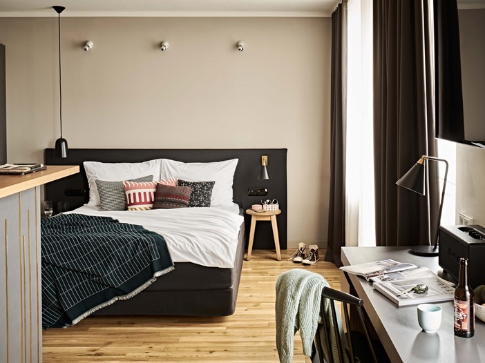 Ein bequem aussehendes Doppelbett mit bunten Kissen, links eine Theke, rechts ein großes Fenster mit weißen und grauen Stores, ein Schreibtisch mit Stuhl an der Wand, darüber ein Flachbildfernseher. 