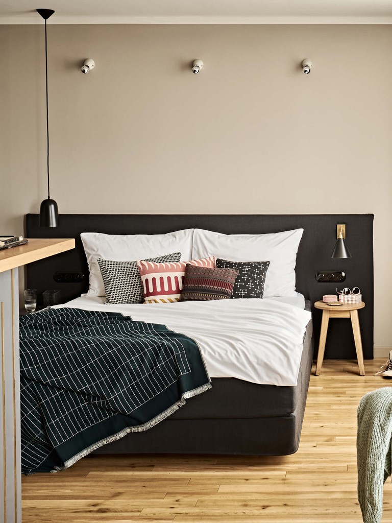 Ein bequem aussehendes Doppelbett mit bunten Kissen, links eine Theke, rechts ein großes Fenster mit weißen und grauen Stores, ein Schreibtisch mit Stuhl an der Wand, darüber ein Flachbildfernseher. 