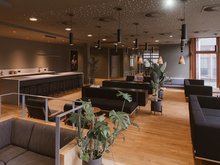 Blick in eine Lounge mit hellen Holzfußboden, modernen Sitzgelegenheiten und Pflanzen. Links befindet sich eine große, offene Küche.