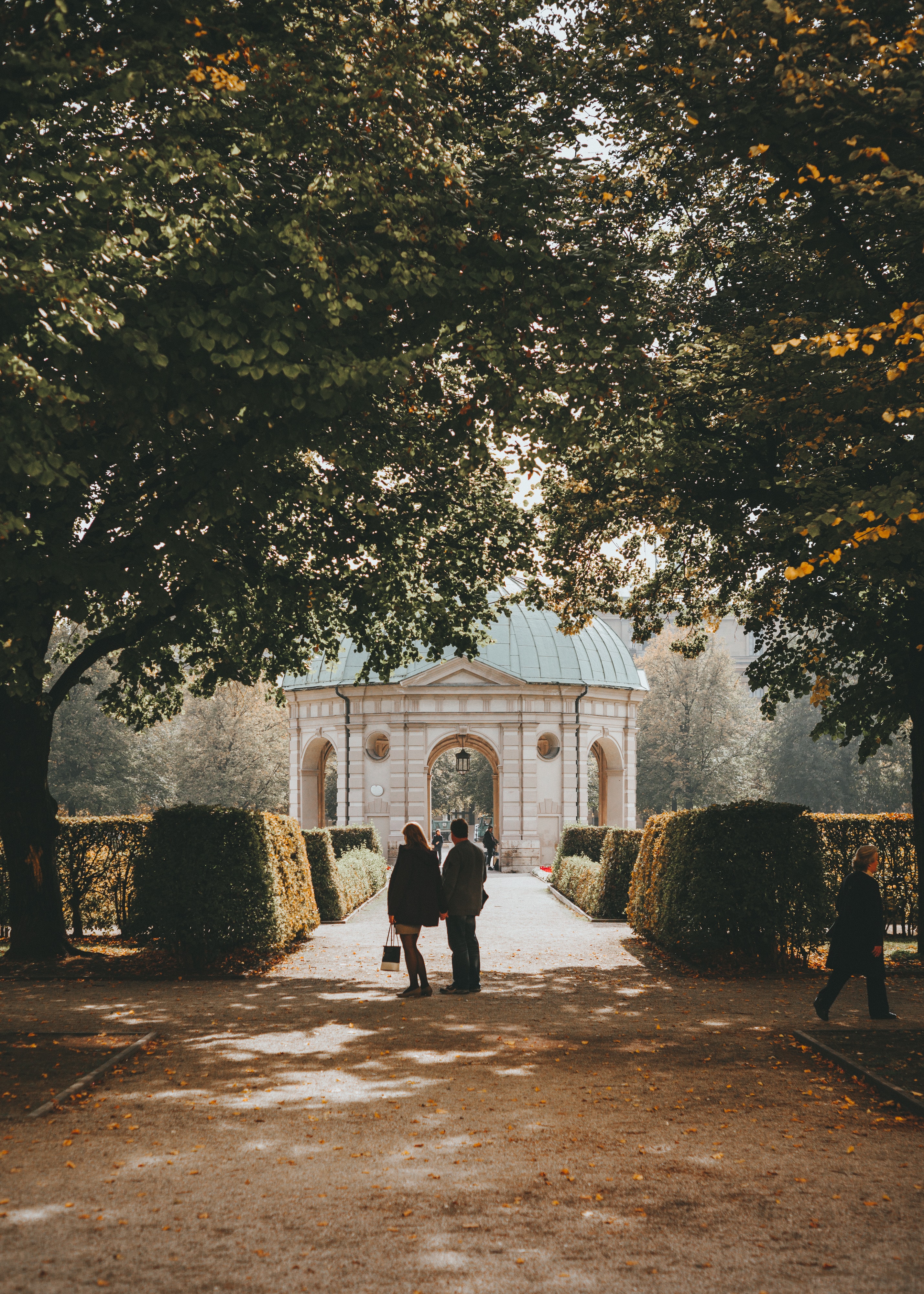 Ein Mann und eine Frau spazieren durch einen herbstlichen öffentlichen Garten, im Hintergrund ein Pavillon aus der Renaissance
