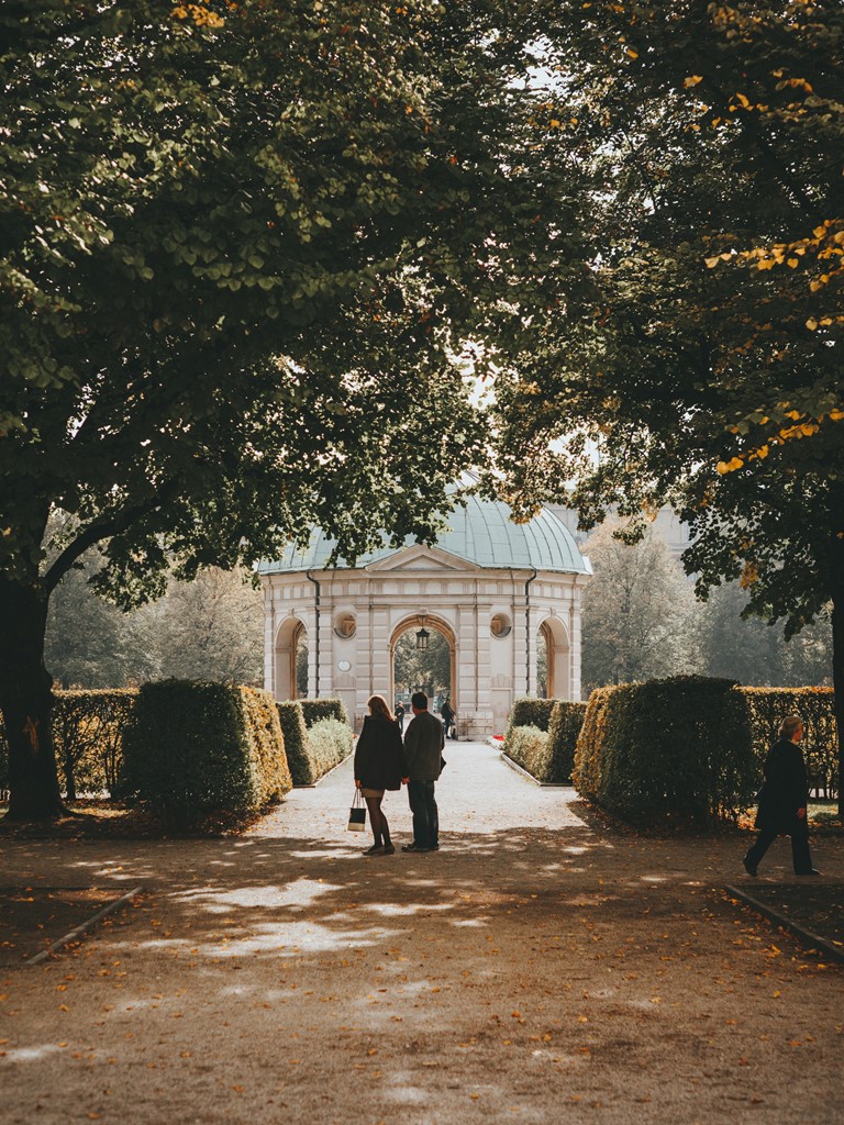 A man and a woman strolling through an autumnal public garden, a Renaissance pavilion in the background 