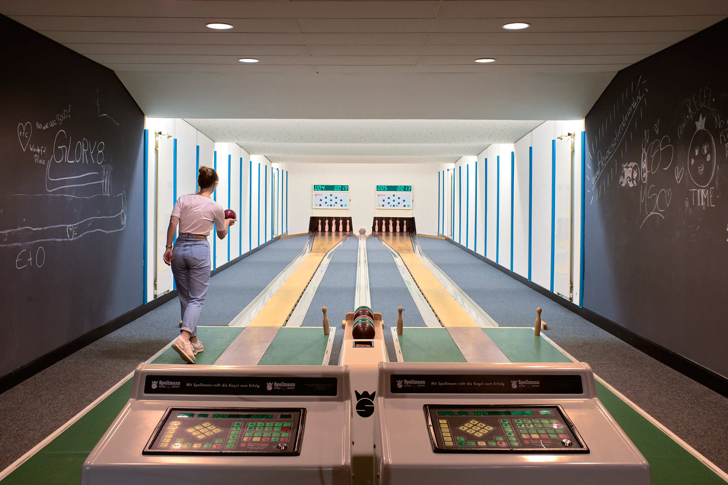 Zwei Kegelbahnen aus den 70er Jahren mit Displays, die Wände sind mit Kreide bemalt, links setzt eine junge Frau in Jeans zum Wurf an. 