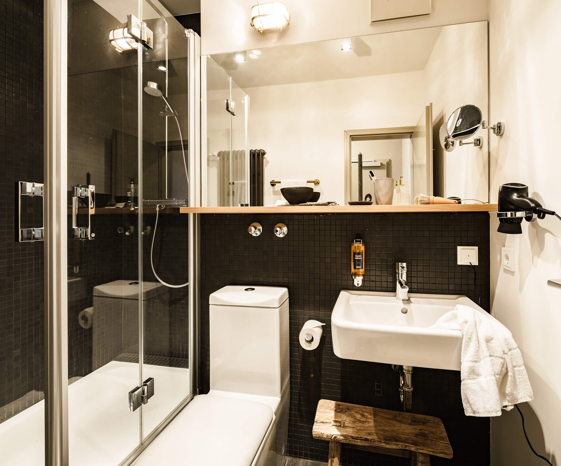A bathroom in modern style, a glass shower cubicle on the left, a large mirror in the middle, a white toilet and washbasin below, and a wooden stool. 