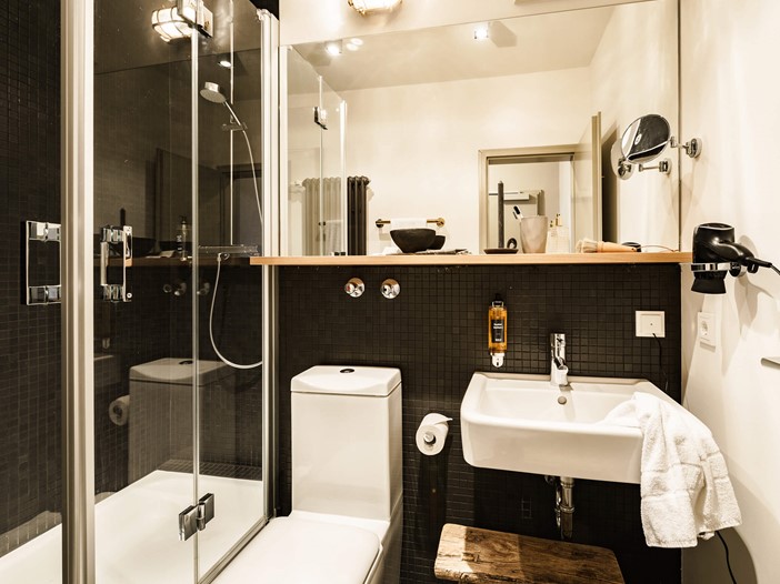 Ein Badezimmer in modernem Style, links eine gläserne Duschkabine, mittig ein großer Spiegel, darunter eine weiße Toilette und ein Waschbecken sowie ein hölzerner Hocker. 