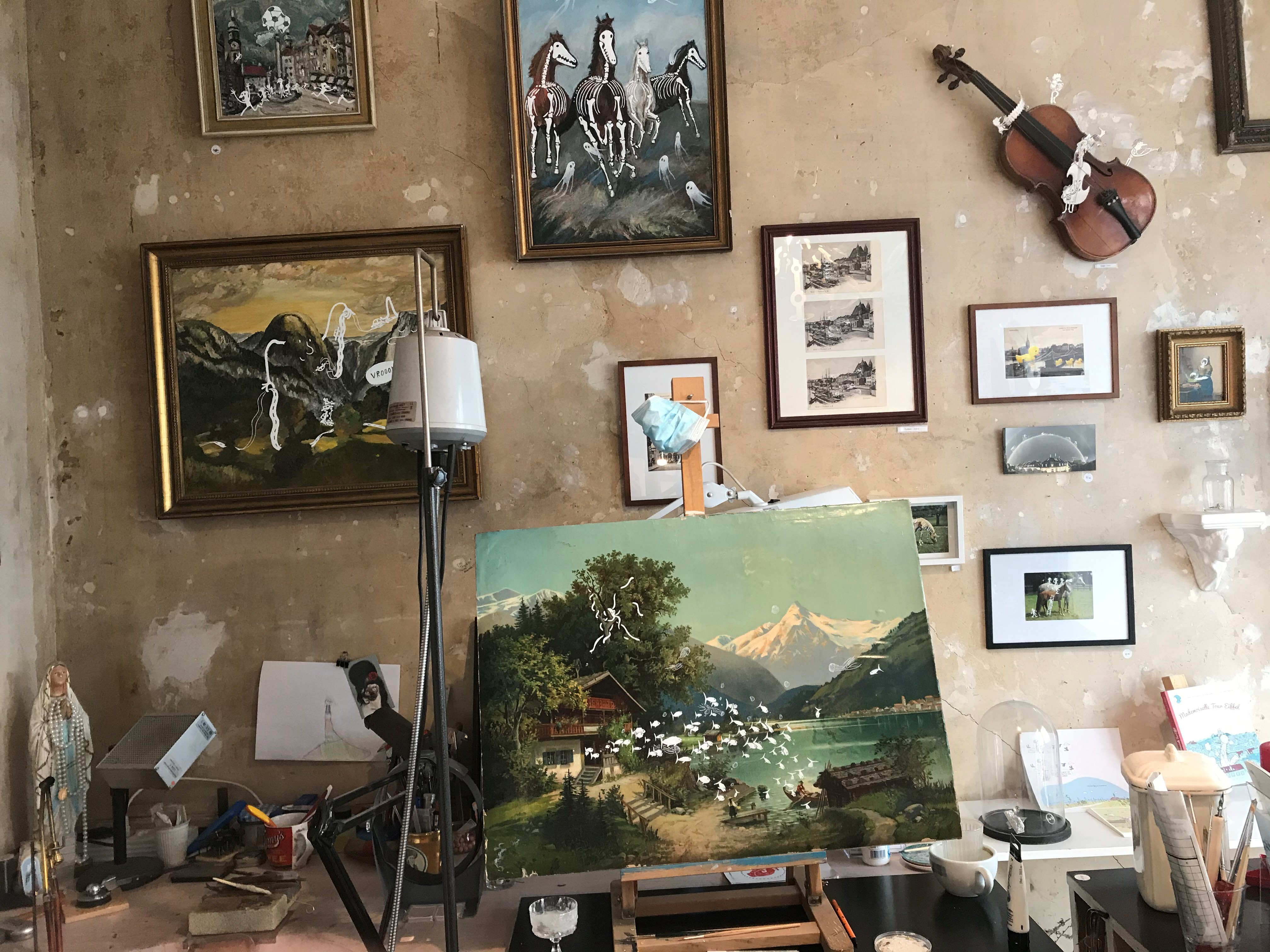 Ein Gemälde auf einer kleinen Tischstaffelei. An der Wand hängen verschiedene andere Gemälde und Fotos. 