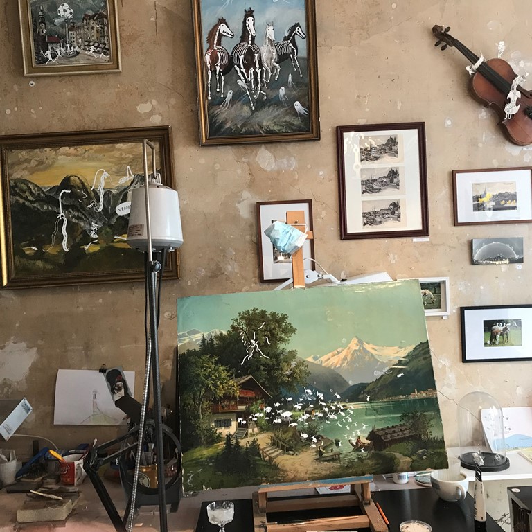 A painting on a small table easel. Various other paintings and photos hang on the wall.