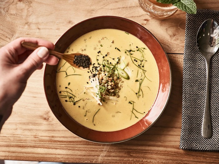A brown, deep plate with cream-coloured soup on a wooden table, next to it a spoon on a textile napkin. A small spoon is used to sprinkle black sesame seeds on the plate. 