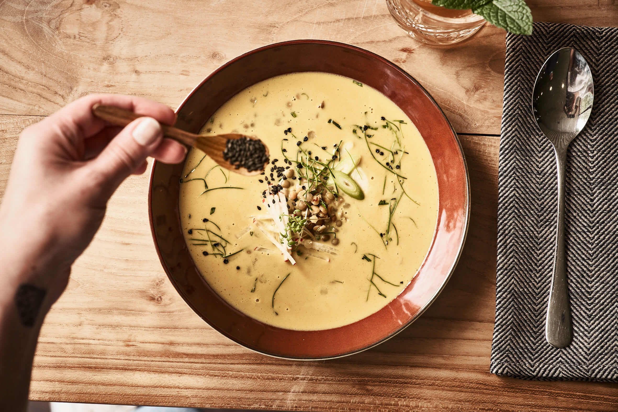 Ein brauner, tiefer Teller mit cremefarbener Suppe auf einem Holztisch, daneben ein Löffel auf einer Serviette. Mit einem kleinen Löffel wird schwarzer Sesam auf den Teller gestreut. 