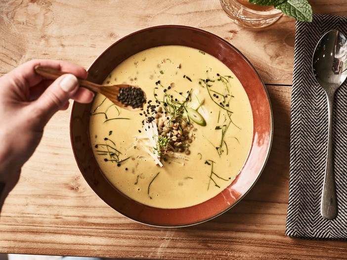 Ein brauner, tiefer Teller mit cremefarbener Suppe auf einem Holztisch, daneben ein Löffel auf einer Serviette. Mit einem kleinen Löffel wird schwarzer Sesam auf den Teller gestreut. 