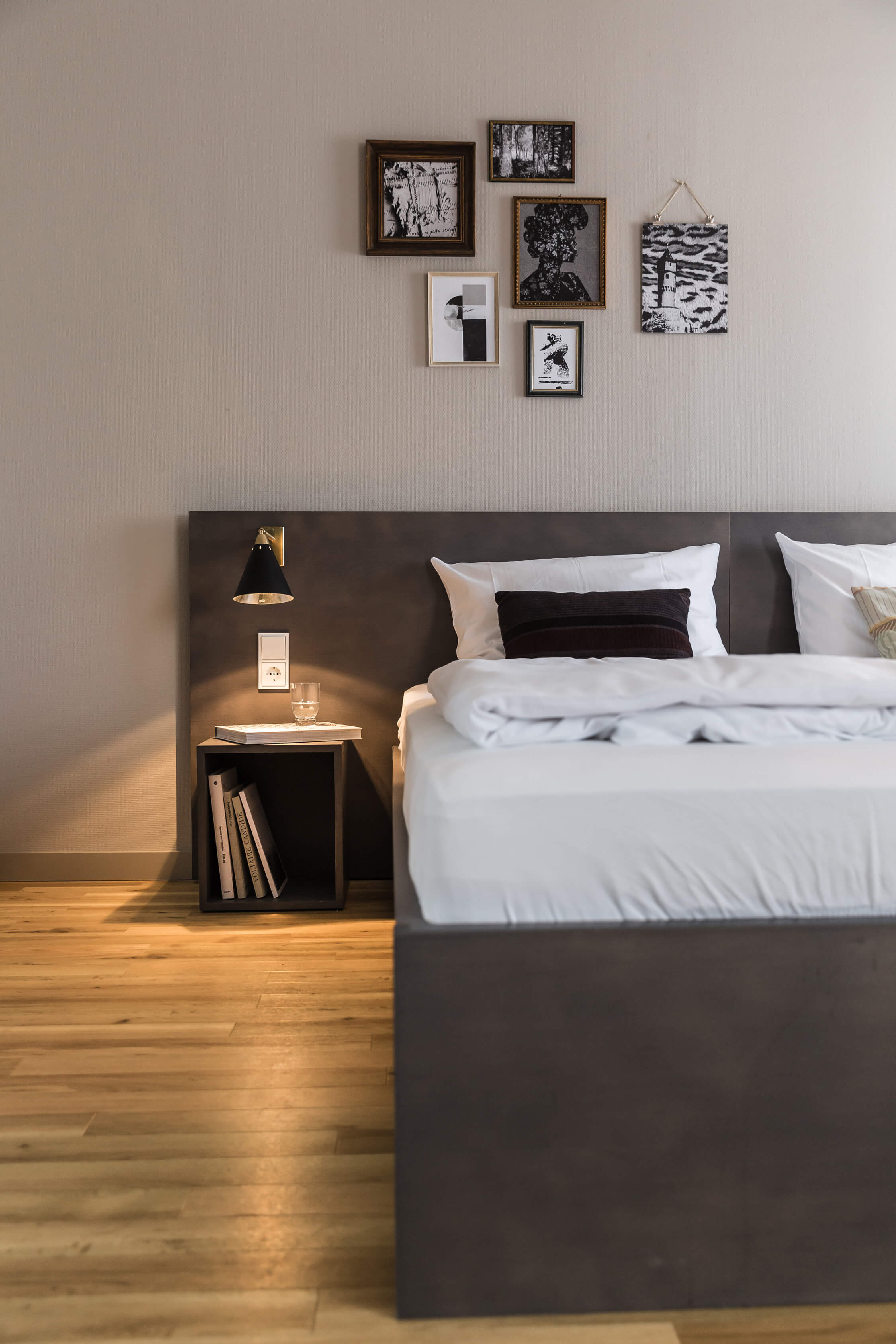 View of a wooden floor and a double bed on the right, next to it a bedside table with a water glass, books and a lamp above it. Modern pictures decorate the wall behind it.