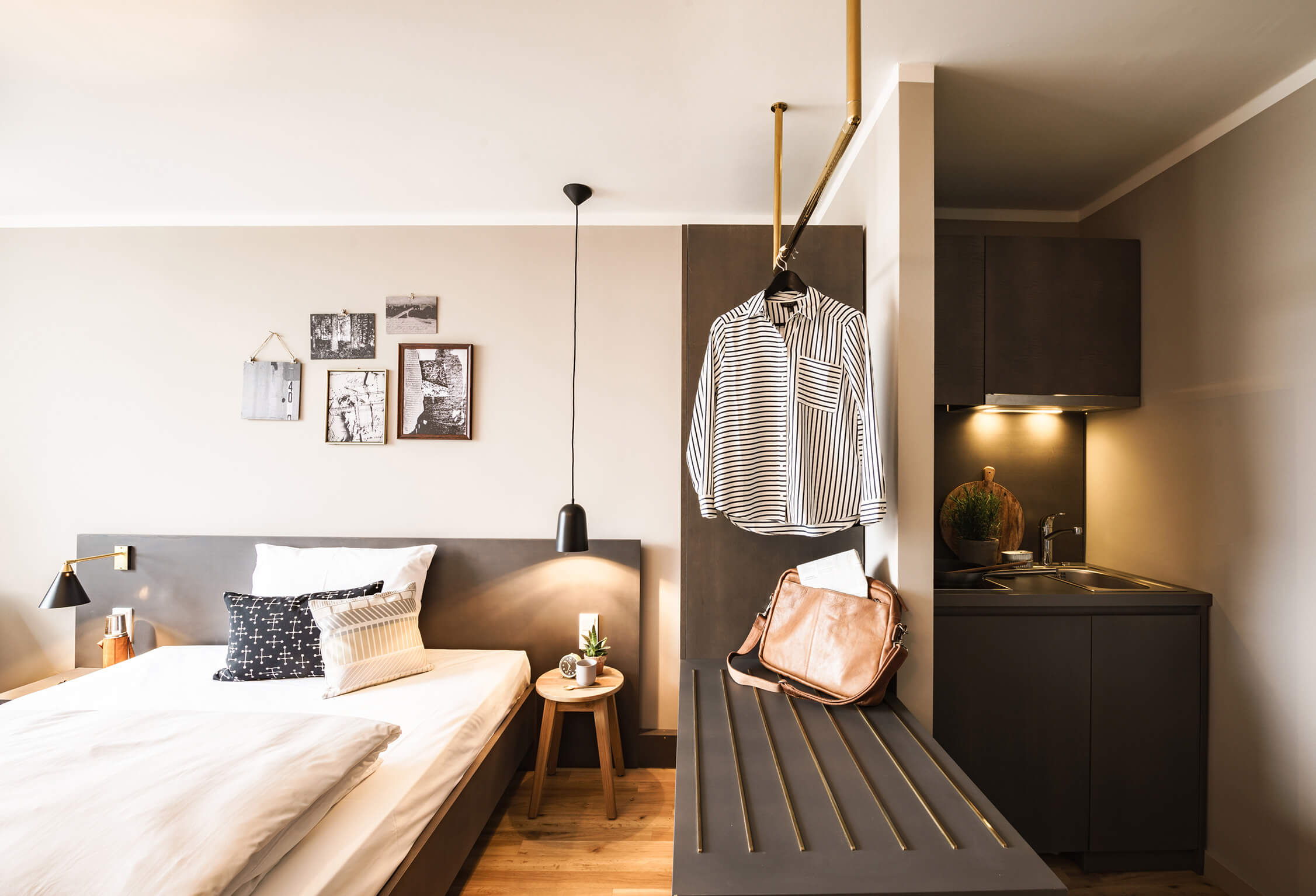 Helles Zimmer mit Holzboden. Links ein bequemes Bett mit Kissen und Bildern an der Wand, mittig ein goldenes Designelement als Garderobe, rechts leicht abgetrennt, eine beleuchtete Kitchenette. 