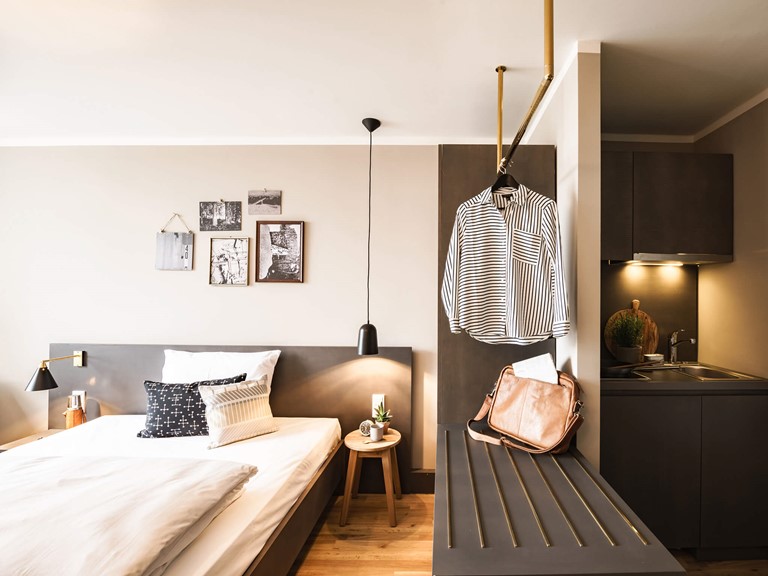 Helles Zimmer mit Holzboden. Links ein bequemes Bett mit Kissen und Bildern an der Wand, mittig ein goldenes Designelement als Garderobe, rechts leicht abgetrennt, eine beleuchtete Kitchenette. 