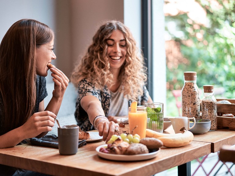 Zwei Frauen sitzen zusammen an einem Tisch und genießen ein reichhaltiges Frühstück mit Blick aus dem Fenster, sie lächeln und unterhalten sich.