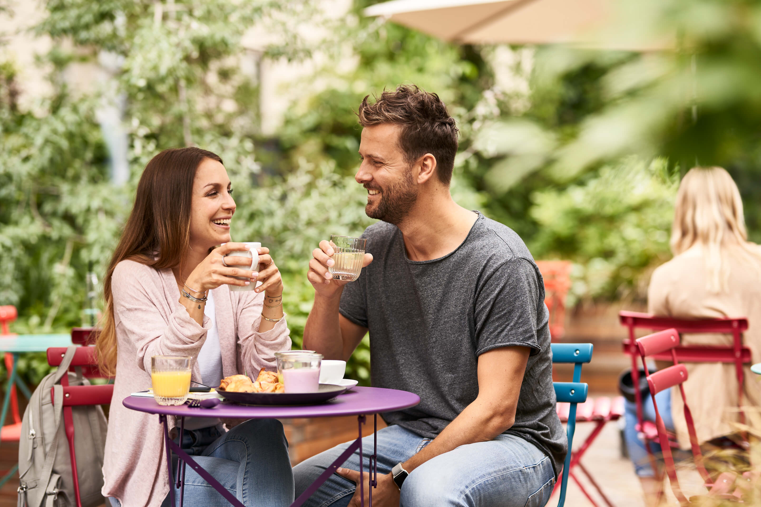 In a leafy courtyard, stylish, colourful chairs with a woman with long brown hair sitting on the left, holding a coffee cup. To her right is a man with brown hair and a beard, they are smiling at each other. 