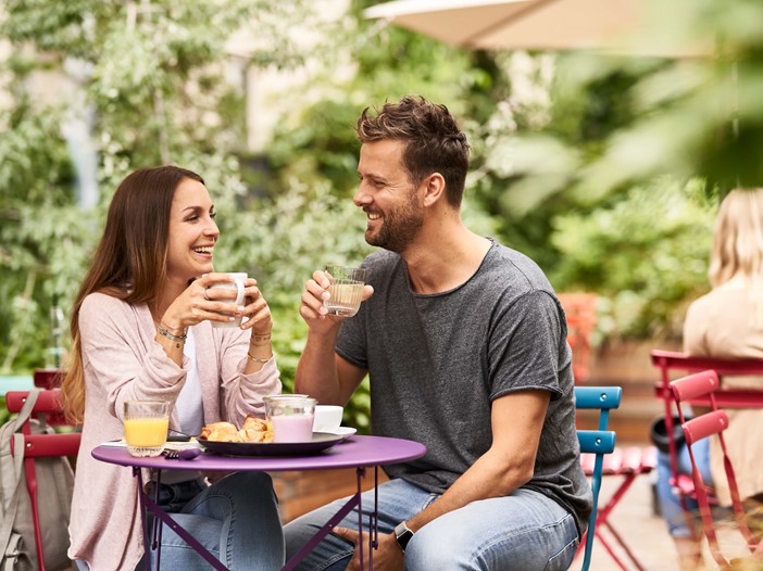 In a leafy courtyard, stylish, colourful chairs with a woman with long brown hair sitting on the left, holding a coffee cup. To her right is a man with brown hair and a beard, they are smiling at each other. 