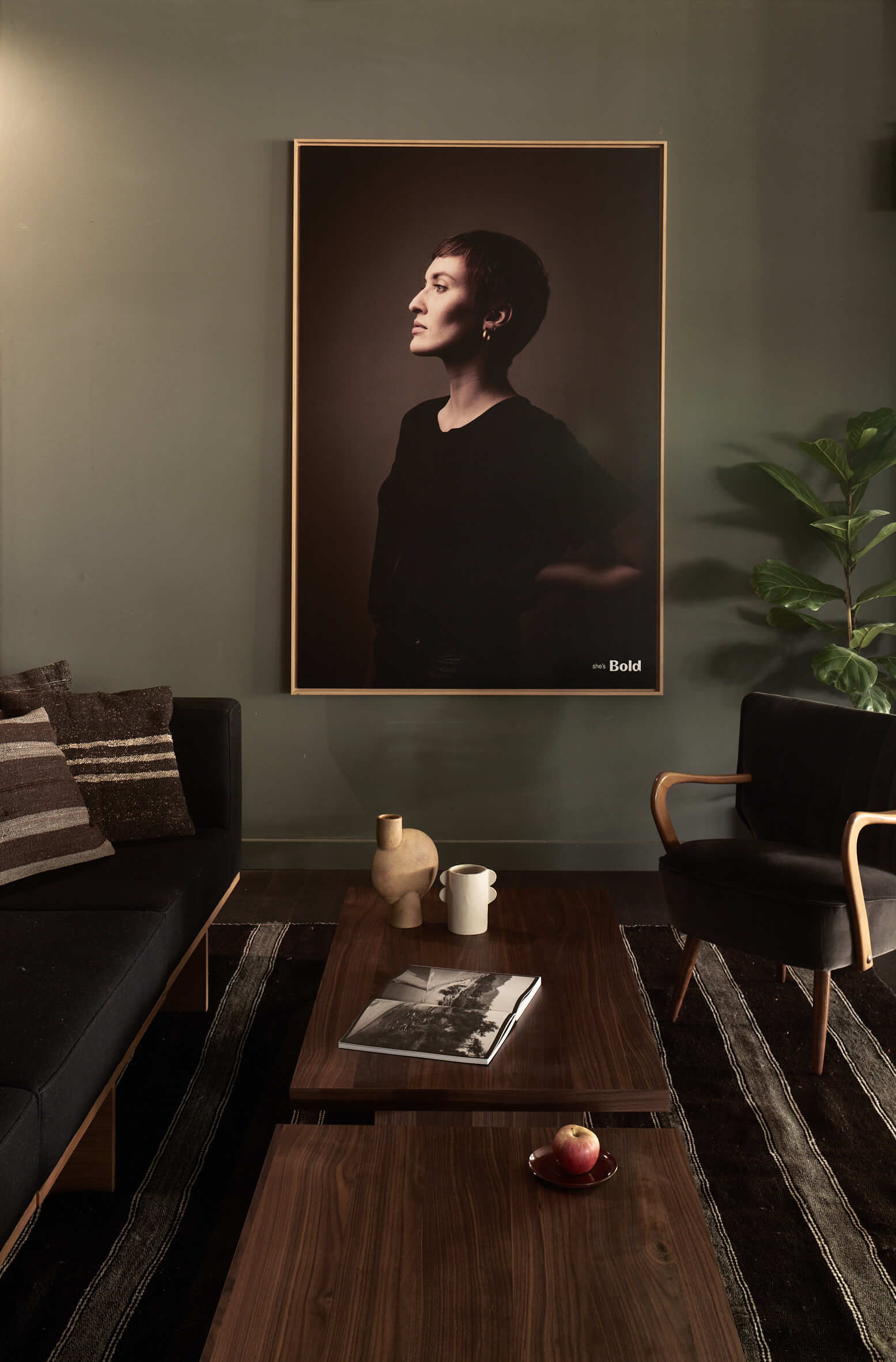 Zentral ein großes, gerahmtes Bild einer kurzhaarigen Frau im Profil. Davor niedrige Holztische auf einem Teppich, daran eine dunkle Couch mit Kissen und ein Sessel.