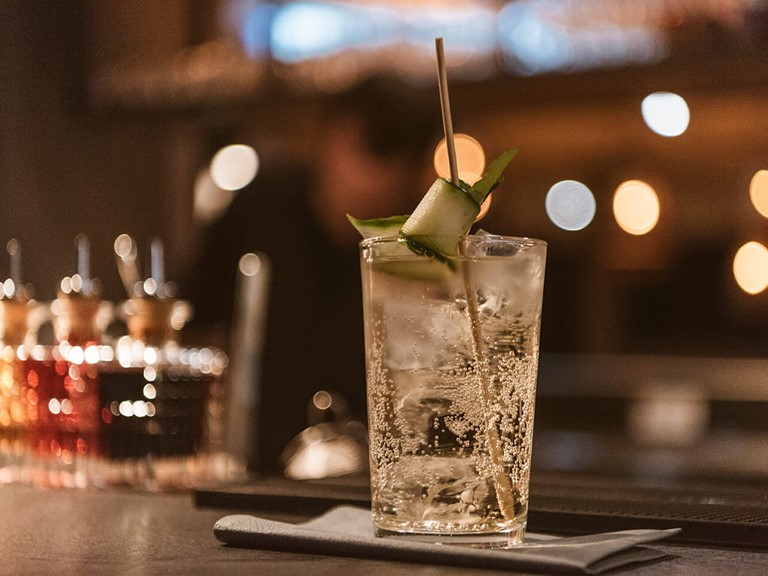 Fokus auf ein erfrischend aussehendes Glas Gin Tonic mit Gurkendeko auf der Theke einer Bar mit abendlicher Beleuchtung. 