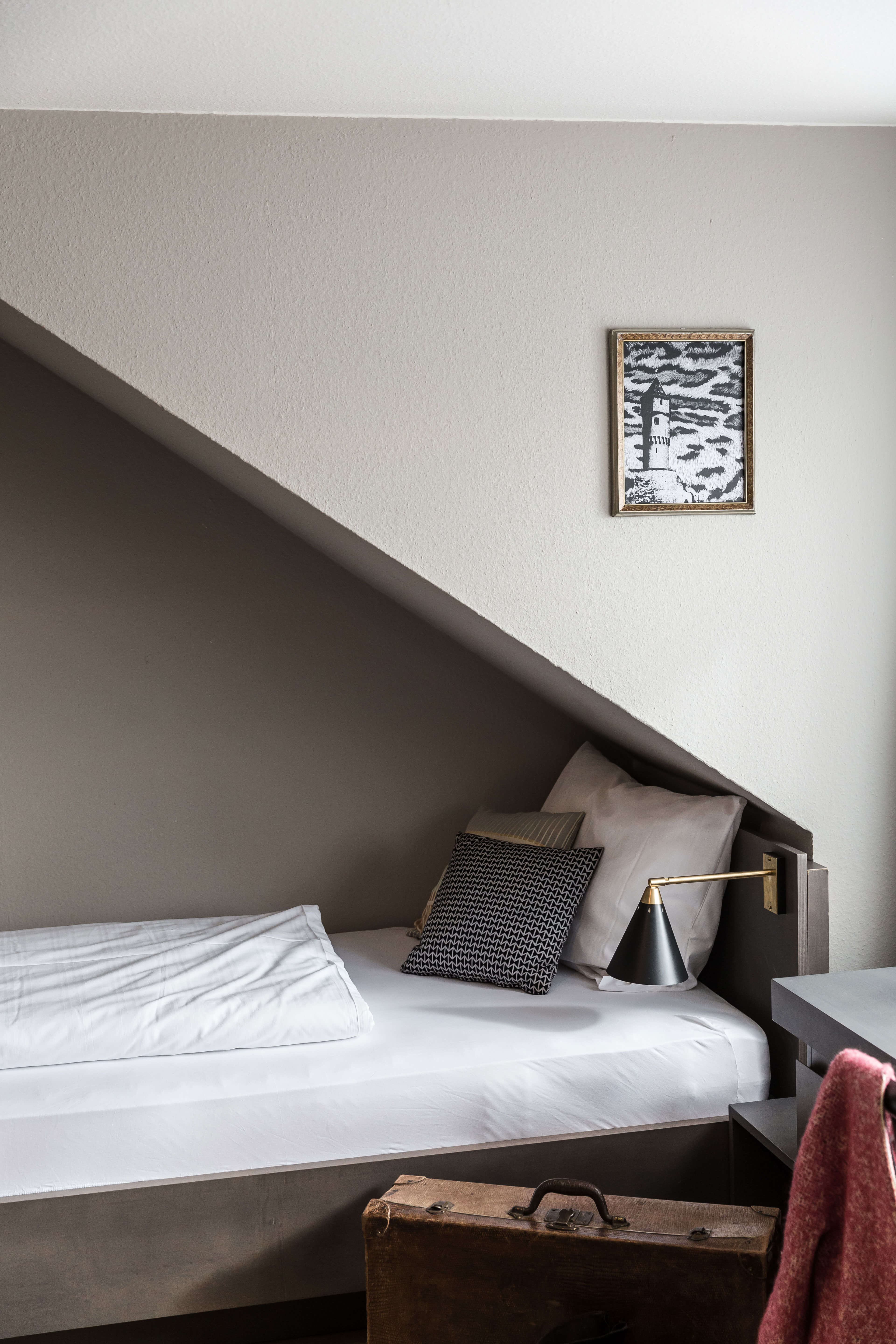 A single bed in the front, with a reading lamp and a travel suitcase in front of it. A golden framed picture hangs on the wall. 