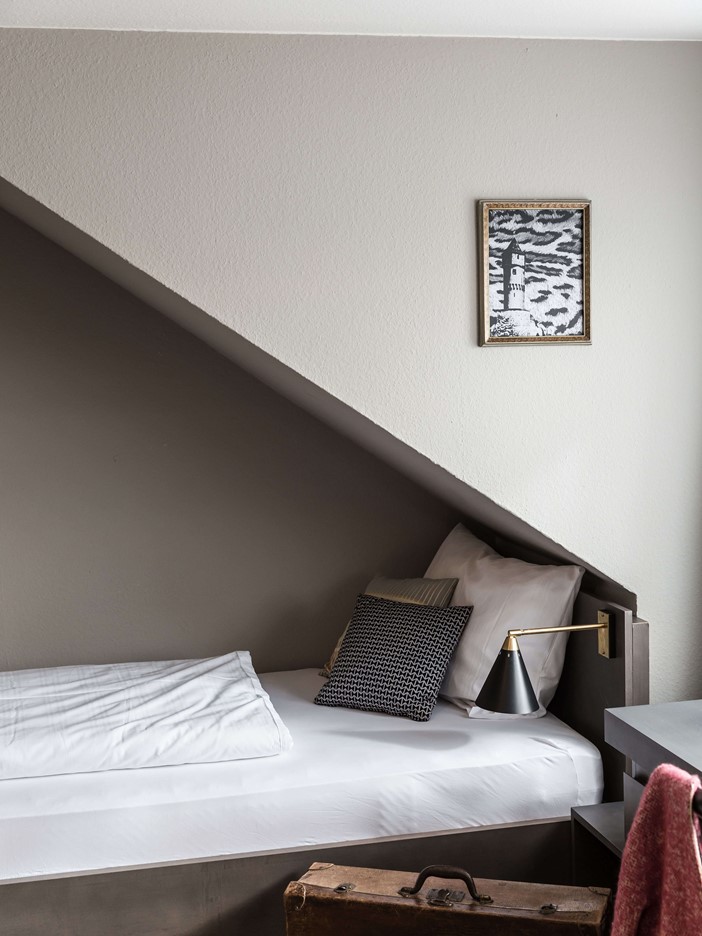 Ein Einzelbett im Anschnitt, mit Leselampe und einem Reisekoffer davor. An der Wand hängt ein golden gerahmtes Bild. 